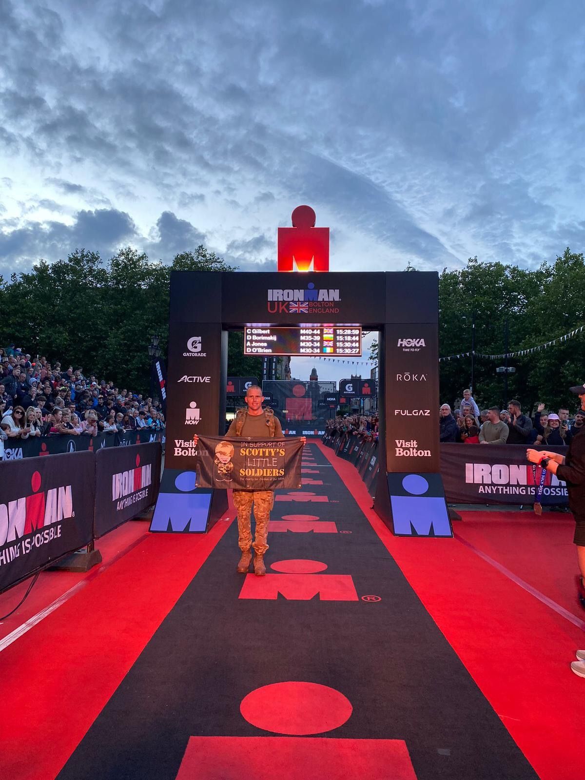 REME vehicle mechanic, Cpl Alan Jones, undertaking his first ever Ironman – the Bolton Ironman Challenge – wearing full military gear for Scotty's Little Soldiers, holds banner at finish line